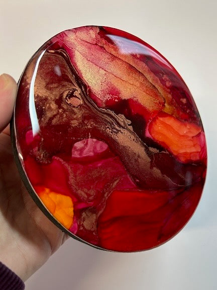 A circle painting of red and orange with copper and gold metallic held by a hand against a neutral background. The rich, fiery colours reflect the phoenix symbolism of rebirth and alchemy and is a encouragement to believe in yourself