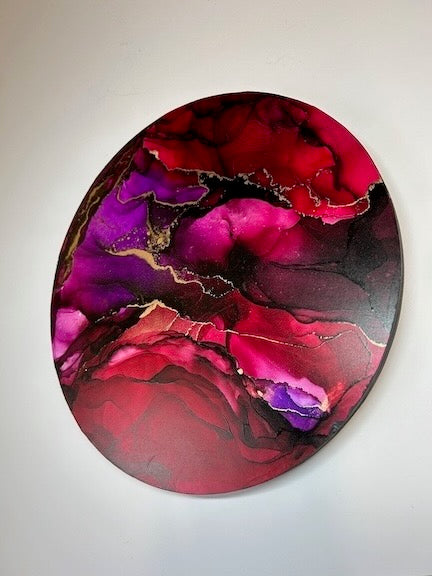 A round painting of an abstract artwork displayed at an incline showing depth and jewel tones - dark purple and red colours. Contemporary painting exploring transformation, resilience and renewal symbolism based on the phoenix fire bird in intuitive mixed media - by Inkandescent Arts.