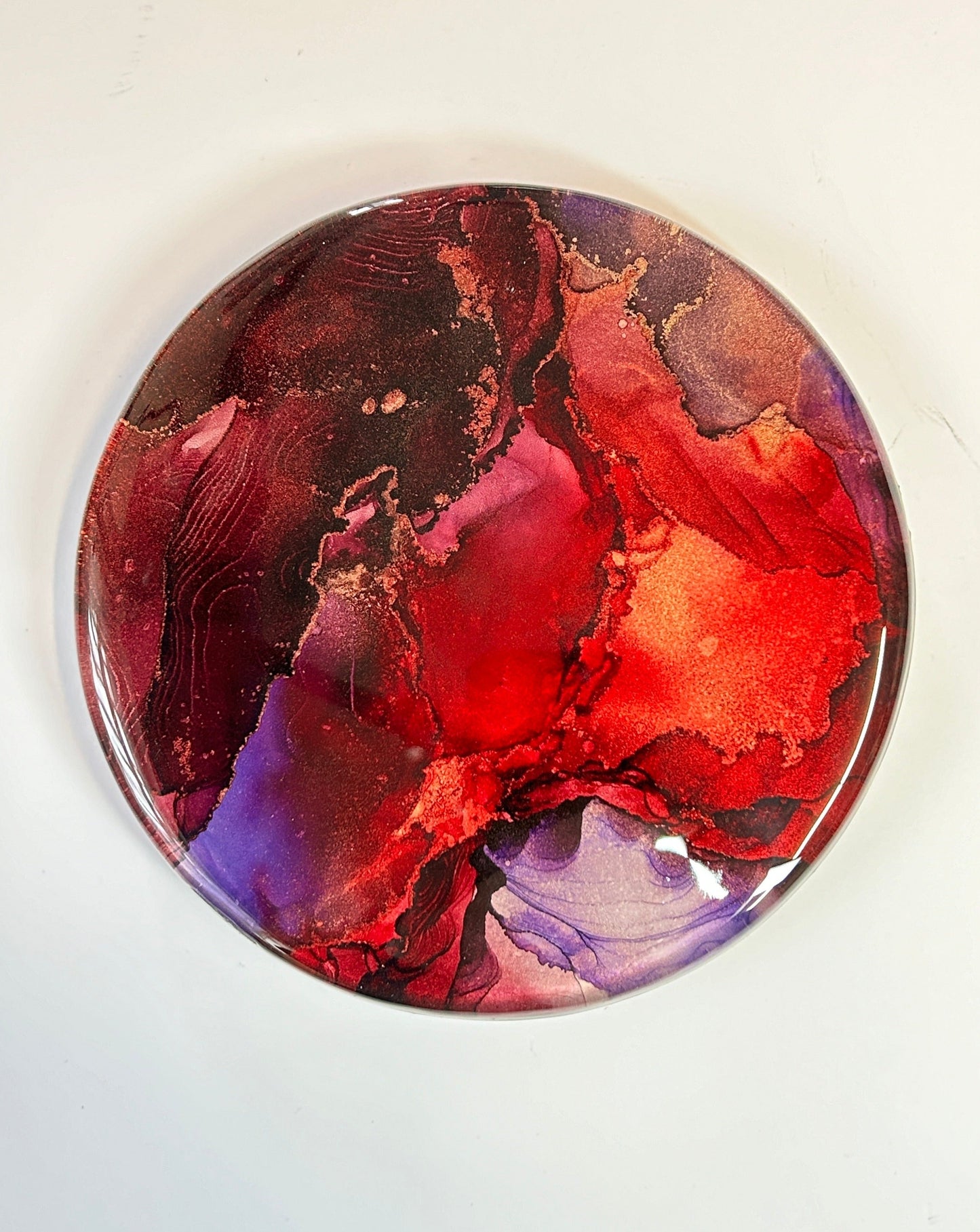 Interior abstract fluid art. Fluid tones of red, violet and maroon flow and overlap in this resin art painting. Copper details appear throughout. 6” circular wall art.