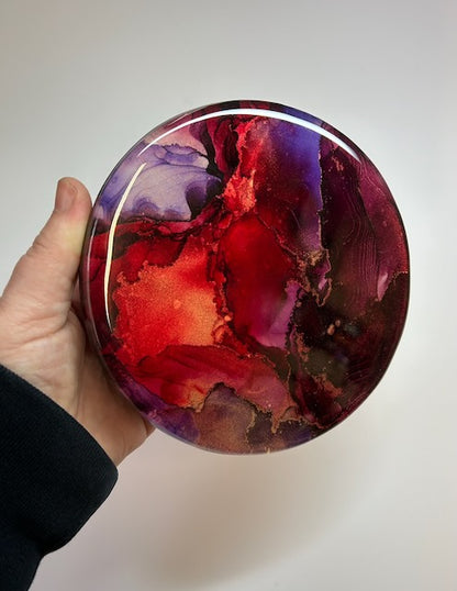 Hand holding round abstract art with bold red and purple colour. Transformative artwork designed to celebrate our triumphs as we rise from the ashes. Renewal symbolism by Inkandescent Arts.