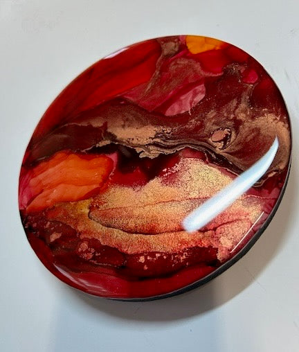 A small, round, abstract painting displayed angled to the left showing shimmering gold detail and depth in scarlet, maroon and orange colours.