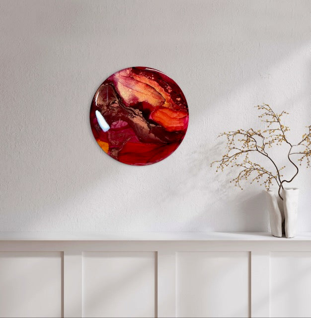 Small wall art in a round design decorates a wall above a radiator cover and vase. A rebirth painting, and transformation symbol in gold, copper, fiery red and warm orange that reflects death - the major arcana card and the Phoenix rising by Inkandescent Arts.