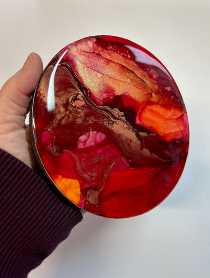 A small round painting of transformation symbolism hand held at an angle, showing spicy warm reds, orange and metallics.
Art based on the Arcana death card and Phoenix symbolism by Inkandescent Arts.